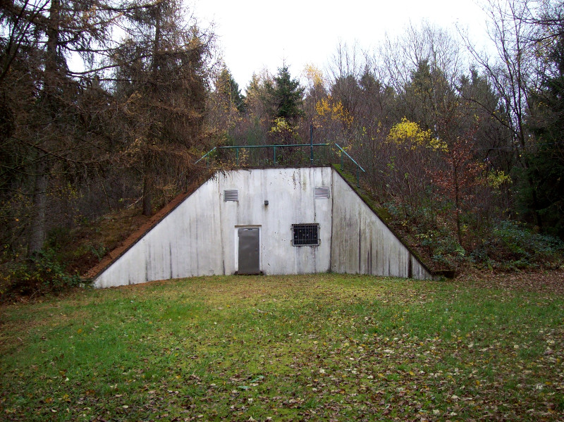 Foto eines trapezförmigen Bunkereingangs auf dessen Dach bereits ein Nadelwald wächst, davor Herbstblätter auf einem Rasen. (c) Simon Klampt